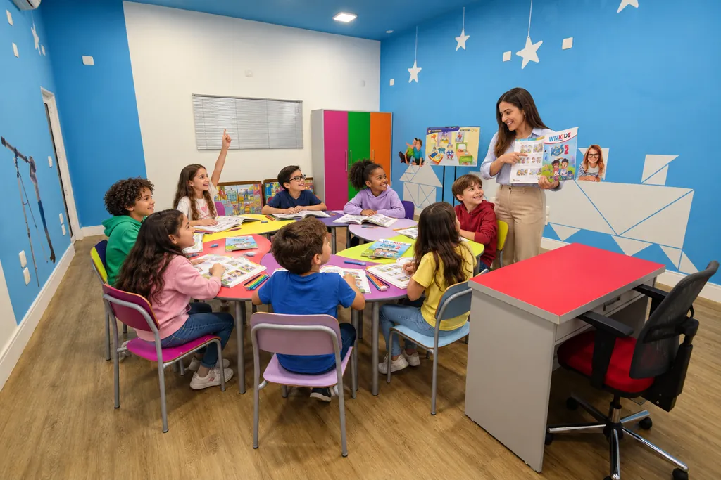 Professora mostrando o livro do curso para a turma Kids em uma sala colorida, com as crianças sentadas em mesas redondas